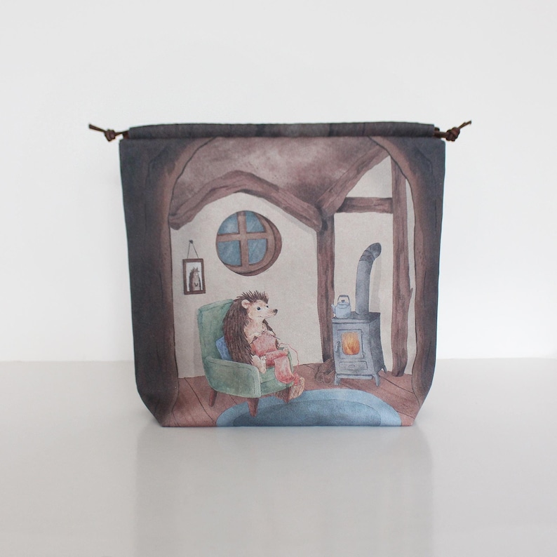K&ouml;nnte beinhalten: Eine braune Canvas-Tragetasche mit einer skurrilen Illustration eines Igels, der in einem gr&uuml;nen Sessel am Kamin sitzt. Der Igel tr&auml;gt einen roten Pullover und h&auml;lt eine Wollkn&auml;uel. Die Illustration spielt in einem gem&uuml;tlichen H&auml;uschen mit einem Fenster und einem Bild an der Wand.