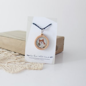 Op de afbeelding: Een houten ketting met een ronde, transparante afbeelding van een rode kater. De hanger hangt aan een blauw koord en is bevestigd aan een kaart met de tekst "the blue rabbit house". Een illustratiewinkel gerund door Eve Vercauteren. Handgemaakt in België.
