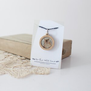 May include: A wooden pendant necklace with a round, light brown wooden frame featuring a printed image of a bear with a white background. The pendant is hanging from a black cord and is displayed on a white card with the text "the blue rabbit house" and "An Illustration Business run by Eva Vercaderen Handmade in Belgium".