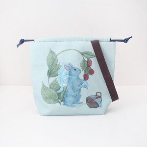 Può includere: Una borsa con coulisse azzurra con un'illustrazione stampata di un coniglio blu con un nastro azzurro intorno al collo, vicino a foglie verdi, lamponi rossi e un piccolo cestino marrone pieno di bacche.