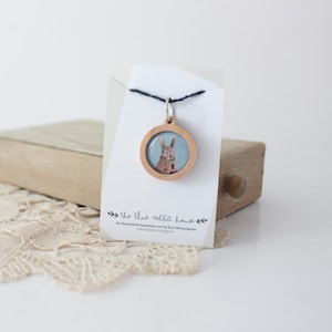 Peut inclure: Un collier avec un pendentif en bois rond sur fond bleu clair représentant une illustration peinte à la main d'un lapin brun. Le pendentif est attaché à un cordon noir et pend d'une carte blanche avec le texte "the blue rabbit house" et "An illustration business run by Eva Vercauteren. Handmade in Belgium."