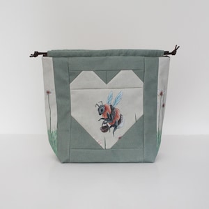 Può includere: Una borsa con coulisse con un design a forma di cuore. La borsa presenta un'illustrazione ad acquerello di un'ape che trasporta un cestino. La borsa è principalmente verde chiaro con un cuore bianco e accenti floreali.