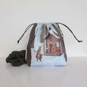 May include: A light blue fabric drawstring bag featuring a printed illustration of a brown rabbit wearing a blue scarf near a wooden cabin in a snowy forest. Dark brown yarn is positioned to the left of the bag, which has brown cords.