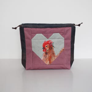 Puede incluir: Una bolsa con cordón con un diseño en forma de corazón que presenta una ilustración de gallo en acuarela. La bolsa tiene un panel central de color malva con un corazón gris, enmarcado por tela negra y gris. El cordón es marrón.