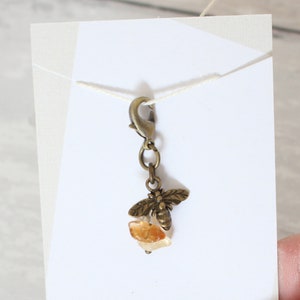Puede incluir: Un amuleto de abeja dorado con un pequeño cristal naranja que cuelga de un cierre de langosta.