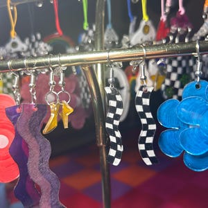 Puede incluir: Una exhibición de pendientes hechos a mano y coloridos. Los pendientes presentan varios diseños, incluyendo patrones ondulados, flores e impresiones de cuadros. Los pendientes están suspendidos de un soporte de pendientes de metal dorado. Los colores incluyen rojo, azul, morado, amarillo y blanco y negro.