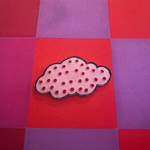 Op de afbeelding: Een roze wolk-vormige broche met rode stippen en een zwarte omtrek ligt op een rood vierkant. De achtergrond is een patchwork van rode, roze en paarse vierkanten. Deze decoratieve broche is een leuk accessoire.