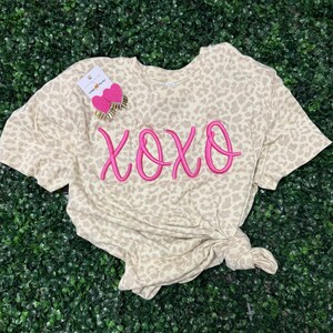 Puede incluir: Una camiseta beige con estampado de leopardo con la palabra "XOXO" bordada en rosa brillante. La camiseta está anudada en la parte inferior y tiene una etiqueta con pendientes de corazón rosa.