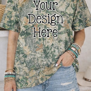 May include: A camouflage-patterned t-shirt with the text "Your Design Here" in a bold, black font. The shirt is paired with light-wash denim shorts. The model is wearing multiple bracelets.