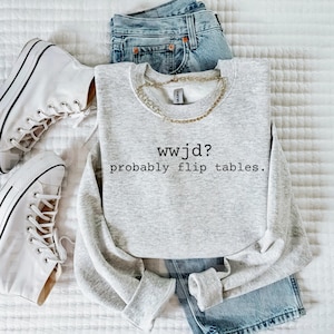 以下が含まれることがあります： 「wwjd？probably flip tables.」の文字が入ったライトグレーのスウェットシャツが、青いジーンズ、ゴールドのチェーン、白いハイカットスニーカーと一緒に写っています。スウェットシャツは白い表面に折りたたまれています。
