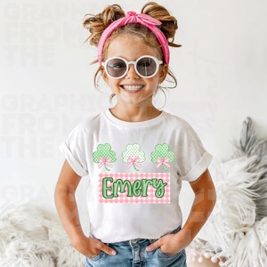 May include: White t-shirt featuring three green shamrocks with pink bows. The shirt has a pink and white gingham design with the name "Emery" in green script. The girl is wearing sunglasses and a pink headband.