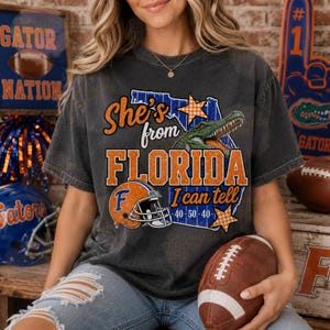 May include: A charcoal gray short-sleeve t-shirt featuring a graphic of the state of Florida in blue, an orange football helmet, a green alligator, and orange stars. The text reads: She's from FLORIDA I can tell 40-50-40.