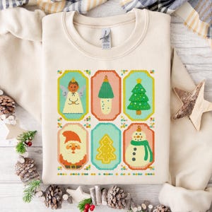 Puede incluir: Sudadera de color crema con un diseño navideño colorido estilo punto de cruz. El diseño incluye imágenes de un ángel, una seta, un árbol de Navidad, Papá Noel, un árbol amarillo y un muñeco de nieve. La sudadera está doblada sobre una superficie de madera blanca.