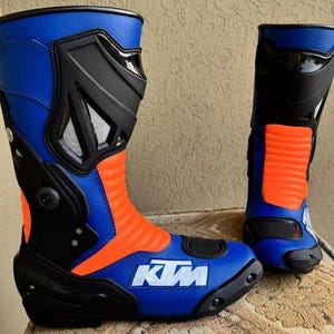 Könnte beinhalten: Paar blaue, orange und schwarze Motorradstiefel. Die Stiefel haben ein blaues Obermaterial, einen orangefarbenen Mittelteil und schwarze Akzente. Der Markenname "KTM" ist in Weiß auf der Spitze des Stiefels sichtbar. Die Stiefel sind auf Schutz und Leistung ausgelegt.