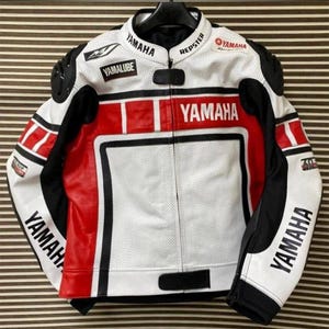 Pode incluir: Casaco de motociclista Yamaha branco, vermelho e preto. O casaco apresenta o logótipo Yamaha no peito, nas mangas e na gola. O casaco tem detalhes em preto nos ombros e nas mangas. O casaco está pendurado num cabide preto.
