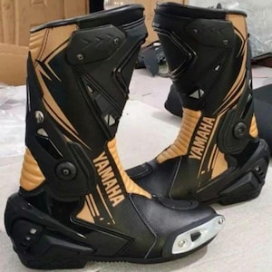 Puede incluir: Par de botas de moto Yamaha negras y beige. Las botas tienen un diseño elegante con el logotipo de Yamaha en letras doradas. Cuentan con elementos de protección y una zona de los dedos reforzada, adecuadas para motociclismo.