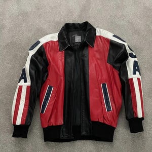 Könnte beinhalten: Vintage Lederjacke mit Colorblock-Design. Die Jacke hat rote, weiße und schwarze Einsätze mit dem Schriftzug "USA" auf den Ärmeln. Sie hat einen schwarzen Kragen, Bündchen und Bund.