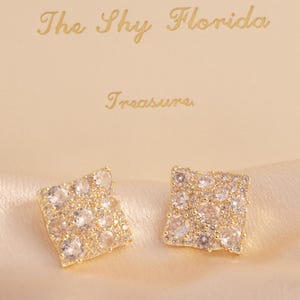 May include: Square-shaped stud earrings with a gold-tone setting and clear, sparkling stones. The earrings are displayed on a satin surface, with a box in the background that reads "The Shy Florida Treasure."