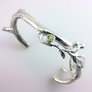 May include: Silver bracelet with a twig design and a green gemstone. The bracelet is open and has a delicate branch with leaves.