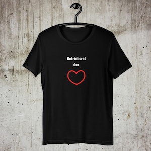 以下が含まれることがあります： 黒いTシャツ。赤いハートの輪郭の上に「Betriebsrat der」の文字がプリントされています。Tシャツは黒いハンガーに掛けられており、背景はニュートラルです。
