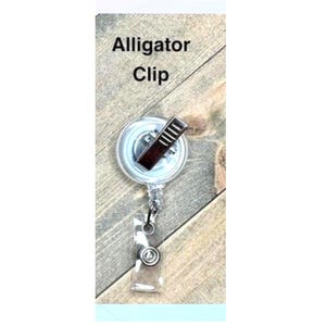 Op de afbeelding: Een doorzichtige, oprolbare badgehouder met een alligatorclip. De haspel heeft een zilverkleurige metalen clip en een transparante plastic band met een druksluiting. De tekst "Alligator Clip" is zwart gedrukt.