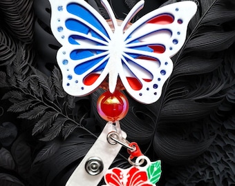 May include: A white butterfly-shaped badge reel with blue and red accents. The badge reel has a red and iridescent bead, a white strap, and a red hibiscus flower charm with green leaves. The background is a dark, stylized floral pattern.