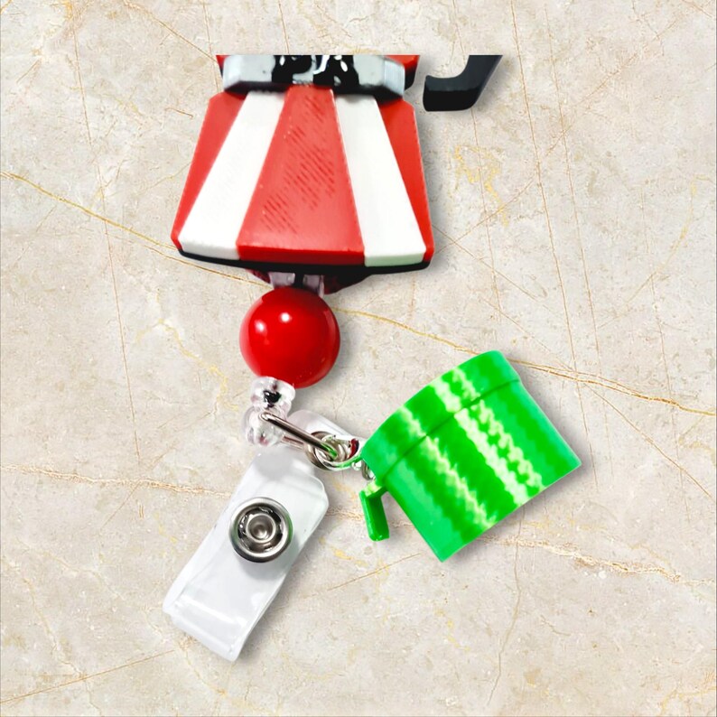 May include: A retractable badge holder with a red and white striped top, a red ball, and a green and white striped accessory. The holder has a white plastic clip and a metal clasp.