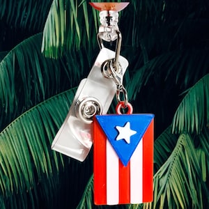 May include: A badge reel featuring the flag of Puerto Rico. The flag is red, white, and blue, with a white star on a blue triangle and white stripes. The reel has a clear plastic clip and a retractable cord.