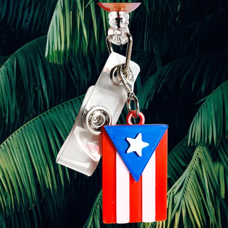 May include: A badge reel featuring the flag of Puerto Rico. The flag is red, white, and blue, with a white star on a blue triangle and white stripes. The reel has a clear plastic clip and a retractable cord.