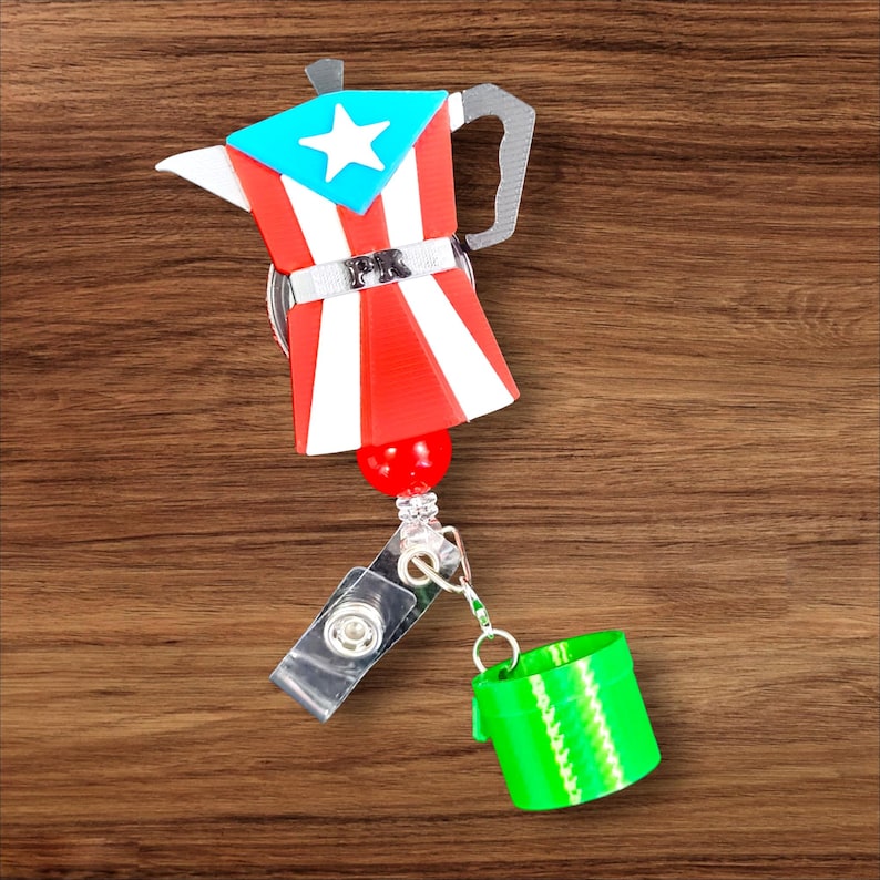 Op de afbeelding: Een badge reel met een koffiepotontwerp in de kleuren van de Puerto Ricaanse vlag: rood, wit en blauw. Het heeft een intrekbaar koord en een groene, cilindrische bevestiging. De koffiepot heeft een zilveren handvat en een rood-wit-blauw ontwerp.