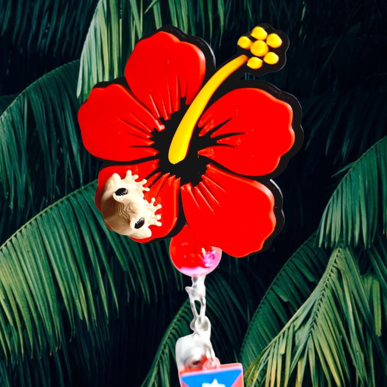 May include: A vibrant red hibiscus flower wind chime with a yellow stamen and black outline. A small, light-colored frog is attached to the flower. The wind chime hangs against a backdrop of green palm leaves.