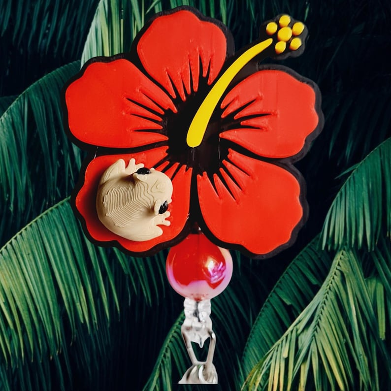 May include: A vibrant red hibiscus flower with a black center, yellow stamen, and a small beige frog attached. A red and pink orb hangs below, suspended by a clear, decorative element. The background features lush green tropical foliage.