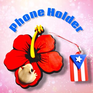 May include: A red hibiscus flower phone holder with a yellow stamen and a small, off-white figure. A small charm with the Puerto Rican flag is attached. The words "Phone Holder" are arched above.