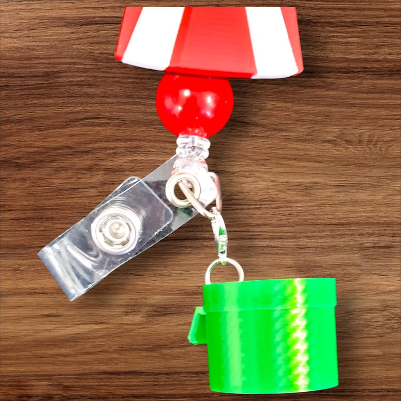 Op de afbeelding: Een rood-wit gestreepte badge reel met een rode bal en een doorzichtige plastic clip. Bevestigd is een groen pijpvormig accessoire met een zigzagpatroon. De reel ligt op een houten oppervlak.