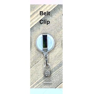 Op de afbeelding: Een heldere, ronde intrekbare badgehouder met een zilveren metalen riemclip. De badgehouder heeft een doorzichtige plastic band en een decoratief zilverkleurig metalen accent. De tekst "Belt Clip" staat op de verpakking.