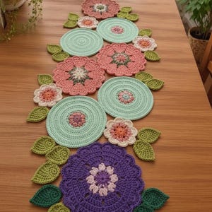 Könnte beinhalten: Ein handgefertigter Häkel-Tischläufer mit einer Reihe kreisförmiger, mintgrüner Matten, durchsetzt mit Blumenmotiven in Rosa-, Lila- und Cremetönen, akzentuiert durch kleine grüne Häkelblätter, die entlang des Läufers angeordnet sind.