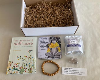 May include: A self-care gift box containing a book titled "the little book of self-care", a coaster with a girl wearing a yellow scarf, a wooden bead bracelet, a shower bomb, and a smoky quartz crystal. The box is filled with brown paper.