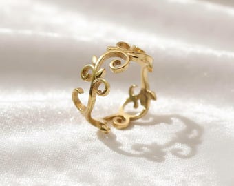 May include: A gold-colored ring with an openwork design featuring swirling floral and leaf motifs. The ring is made of a shiny, metallic material and is set against a white, textured background. The ring's intricate details and delicate curves are highlighted by the light.