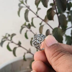 May include: A silver ring featuring an openwork design of swirling patterns. The ring is held up against a backdrop of green and purple foliage.