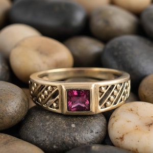 Puede incluir: Anillo dorado con una gema rosa oscuro cuadrada en el centro. La banda presenta un patrón decorativo de líneas diagonales y pequeños acentos redondos. El anillo está colocado sobre un fondo de piedras lisas y multicolores.