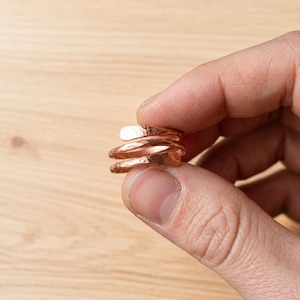 May include: A copper ring featuring a bypass design with a hammered, textured surface. The metal has a warm, metallic reddish-brown finish, and the band wraps around itself in a spiral pattern, creating a distinct layered appearance.