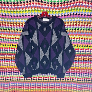 May include: A black, purple, and gray geometric pattern sweater. The sweater has a crew neck and long sleeves. The pattern consists of diamond shapes and lines. The sweater is hanging on a red hanger against a colorful background.