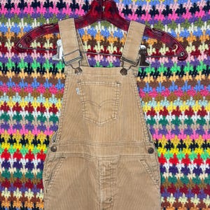 May include: Tan corduroy overalls with adjustable straps and a front pocket. The overalls feature metal hardware and a Levi's brand label. The garment is hanging on a red hanger against a colorful, patterned background.