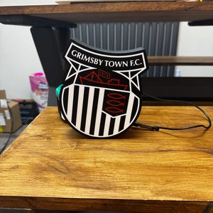 May include: A black and white Grimsby Town F.C. emblem, featuring the team's name and a ship design, sits on a wooden surface. The emblem has a shield shape with vertical stripes and a red ship illustration. A black cable extends from the emblem.