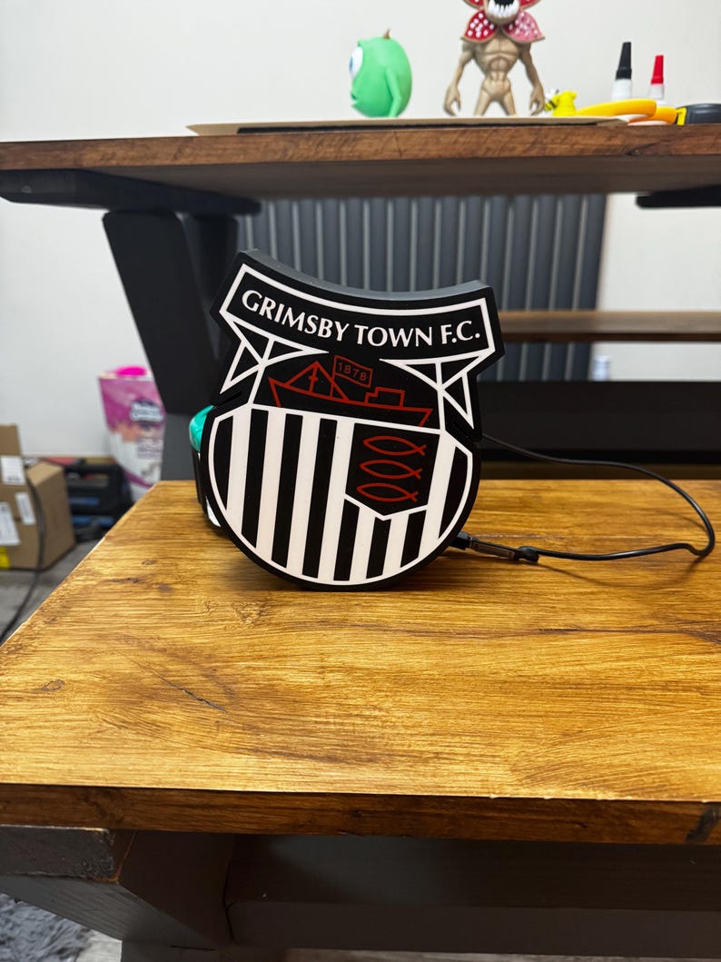 May include: A black and white Grimsby Town F.C. emblem, featuring the team's name and a ship design, sits on a wooden surface. The emblem has a shield shape with vertical stripes and a red ship illustration. A black cable extends from the emblem.