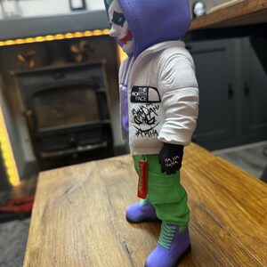 May include: A figurine of the Joker character, wearing a white jacket with "The North Face" logo, green pants, and purple boots. The Joker has green hair, white face paint, and red lipstick. The figurine is standing on a wooden surface.