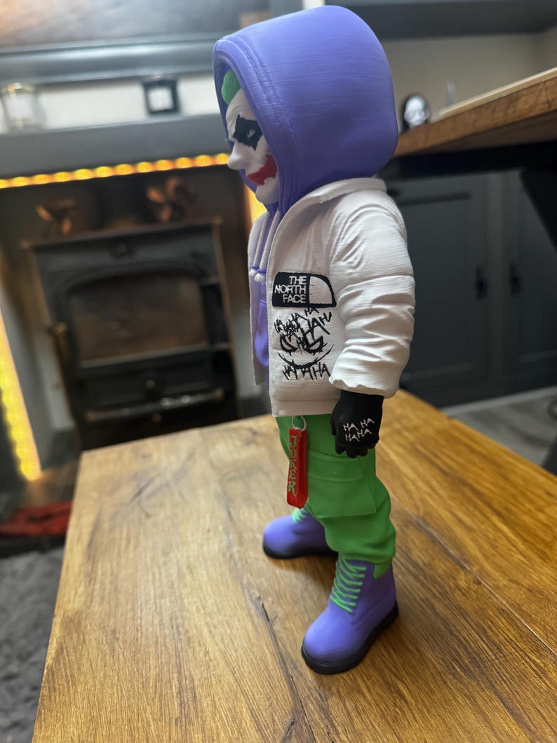 May include: A figurine of the Joker character, wearing a white jacket with "The North Face" logo, green pants, and purple boots. The Joker has green hair, white face paint, and red lipstick. The figurine is standing on a wooden surface.