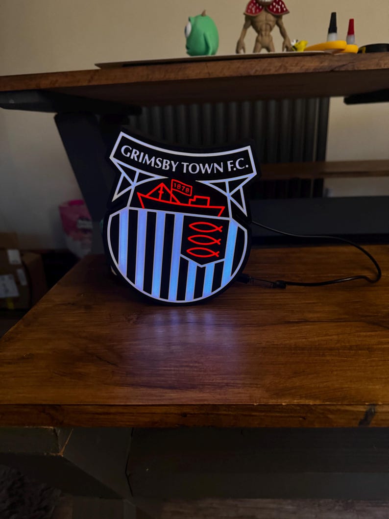 May include: A lighted Grimsby Town F.C. emblem, featuring a shield design with the team's name and a ship illustration. The emblem has a black and white striped background with blue neon lighting. The emblem is on a wooden surface.