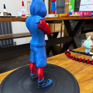 May include: A blue figurine of a character with a hooded sweatshirt, shorts, and sneakers. The figure has red arms and legs. The sneakers are blue and black. The figure is standing on a black circular base.