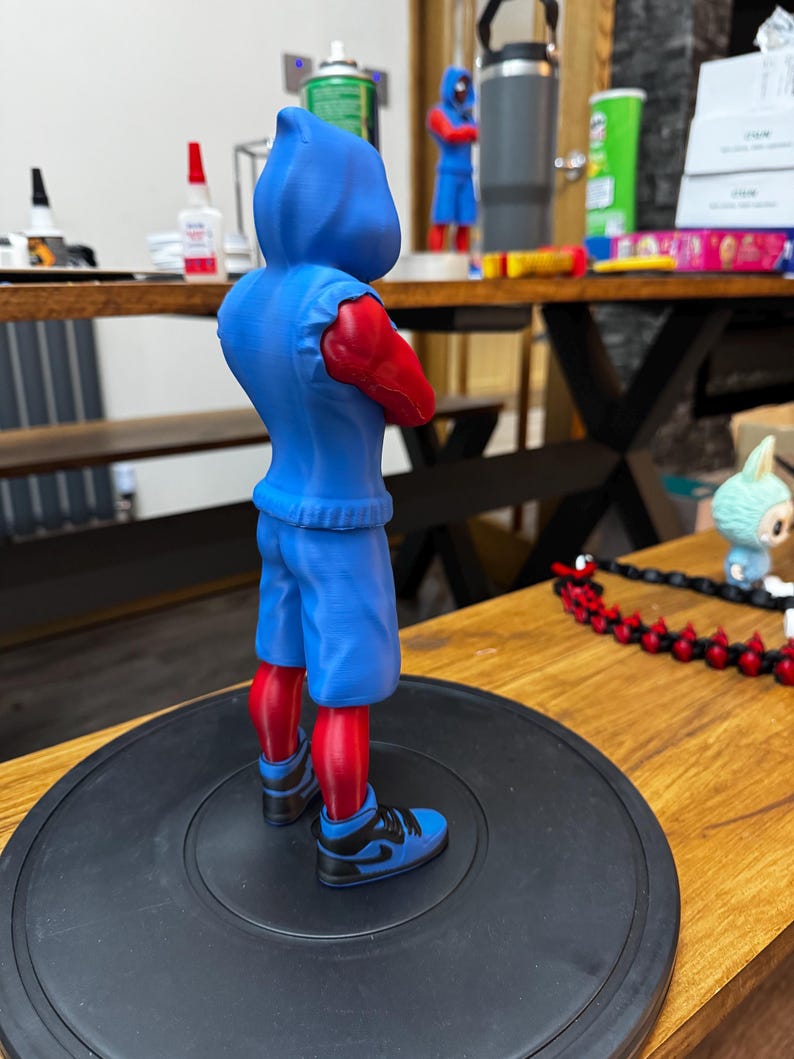 May include: A blue figurine of a character with a hooded sweatshirt, shorts, and sneakers. The figure has red arms and legs. The sneakers are blue and black. The figure is standing on a black circular base.